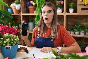 Poster - Hispanic young woman working at florist shop showing smartphone screen scared and amazed with open mouth for surprise, disbelief face