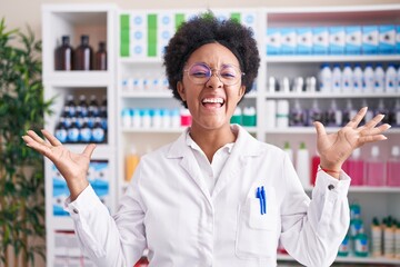 Wall Mural - Beautiful african woman with curly hair working at pharmacy drugstore celebrating victory with happy smile and winner expression with raised hands