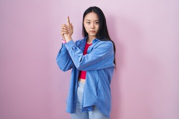 Poster - Young asian woman standing over pink background holding symbolic gun with hand gesture, playing killing shooting weapons, angry face