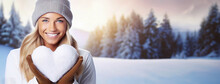 Happy woman holds a snowy heart in winter clothes. Smiling girl face close-up looking at camera. Valentine's Day or winter lover. Snowdrifts and forest landscape background. Panorama, copy space.