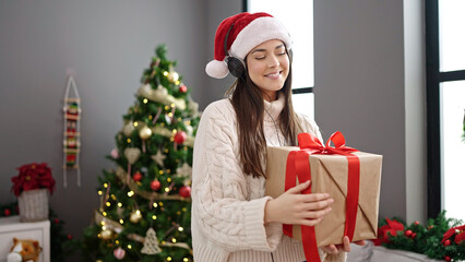 Poster - Young beautiful hispanic woman standing by christmas tree holding gift dancing at home