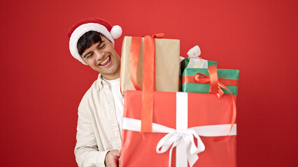 Wall Mural - Young hispanic man wearing christmas hat holding gifts over isolated red background