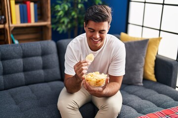 Sticker - Young hispanic man eating chips potatoes sitting on sofa at home