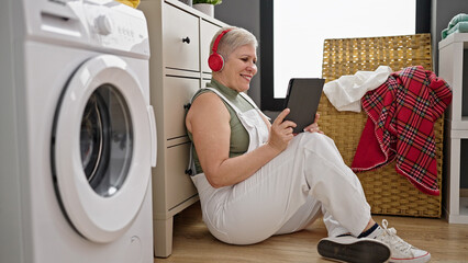 Wall Mural - Middle age grey-haired woman watching video on touchpad waiting for washing machine at laundry room