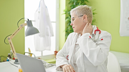 Wall Mural - Middle age grey-haired woman doctor tired stretching neck working at clinic