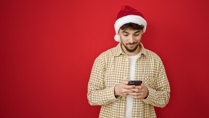 Wall Mural - Young arab man wearing christmas hat using smartphone over isolated red background