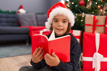 Poster - Adorable hispanic boy reading book sitting on floor by christmas tree at home