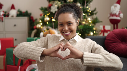 Poster - African american woman doing heart gesture sitting on sofa by christmas tree at home