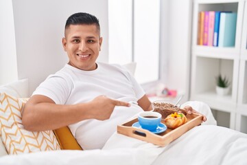 Canvas Print - Young hispanic man eating breakfast in the bed smiling happy pointing with hand and finger