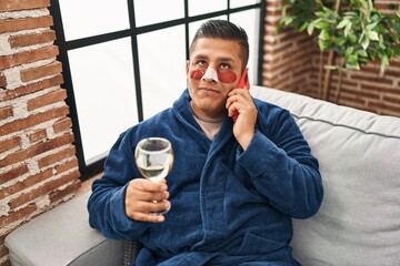 Canvas Print - Hispanic young man wearing bathrobe and eye bags patches drinking wine speaking on the phone smiling looking to the side and staring away thinking.