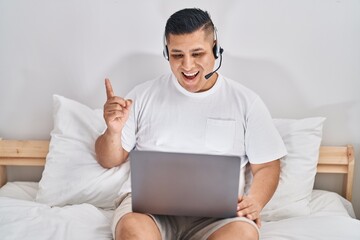 Wall Mural - Hispanic young man using computer laptop on the bed smiling happy pointing with hand and finger to the side