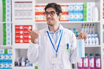 Wall Mural - Young hispanic man working at pharmacy drugstore holding safety mask pointing thumb up to the side smiling happy with open mouth