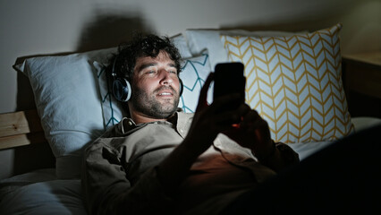 Wall Mural - Young hispanic man watching video on smartphone lying on bed at bedroom