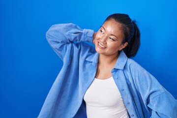 Wall Mural - Asian young woman standing over blue background smiling confident touching hair with hand up gesture, posing attractive and fashionable