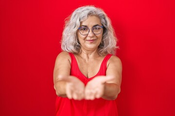 Poster - Middle age woman with grey hair standing over red background smiling with hands palms together receiving or giving gesture. hold and protection