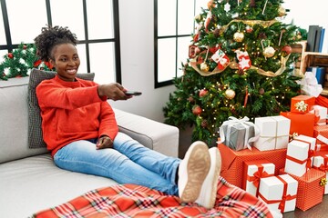 Wall Mural - African american woman watching television sitting on sofa by christmas tree at home