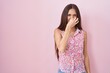 Young hispanic woman with long hair standing over pink background smelling something stinky and disgusting, intolerable smell, holding breath with fingers on nose. bad smell