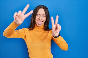 Canvas Print - Young brunette woman standing over blue background smiling with tongue out showing fingers of both hands doing victory sign. number two.