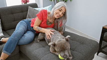 Poster - Middle age grey-haired woman with dog smiling confident lying on sofa playing at home