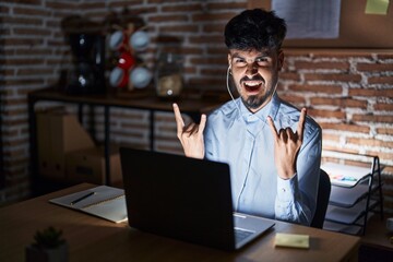 Canvas Print - Young hispanic man with beard working at the office at night shouting with crazy expression doing rock symbol with hands up. music star. heavy concept.