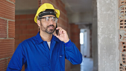 Canvas Print - Young hispanic man worker wearing hardhat talking on smartphone at construction site