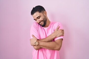 Sticker - Hispanic young man standing over pink background hugging oneself happy and positive, smiling confident. self love and self care