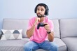 © Krakenimages.com - Hispanic young man playing video game holding controller sitting on the sofa with hand on chin thinking about question, pensive expression. smiling with thoughtful face. doubt concept.
