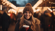 Young woman holding a paper cup with hot drink on Christmas city fair, smiling looking at camera, outdoors Christmas market, European winter holidays