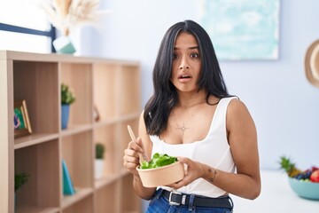 Sticker - Brunette woman eating a salad at home in shock face, looking skeptical and sarcastic, surprised with open mouth