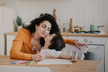 Little curly boy kissing mom at home sitting at desk at kitchen drawing by colour pencils. Joyful young mother eyes closed cuddling with cute son at home. Motherhood, cute family moments.