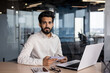 © Tetiana - Portrait of an Indian young businessman working in the office at a table with a laptop, holding a notebook, looking seriously at the camera