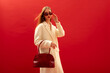Fashionable confident woman wearing trendy white boucle midi coat, stylish sunglasses, holding faux crocodile leather bag, handbag, posing on red background.  Copy, empty space for text