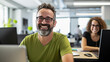 © MP Studio - Cheerful man with glasses and a green t-shirt is smiling at the camera, seated in a modern office with co-workers and computers in the background.