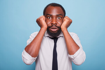 Over a blue background, a nervous entrepreneur with open eyes holds his face in his closed palms. Portrait of a fearful African-American businessman posing scared with his fists over his cheeks.