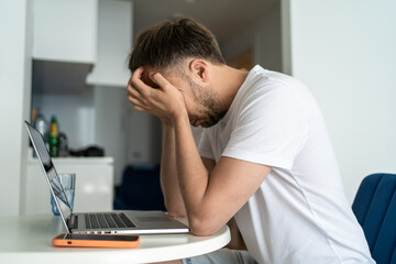 Wall Mural - Overworked man holding head by hands sitting at table working on laptop online distance job from remote home office. Stressed person, middle age crisis concept.