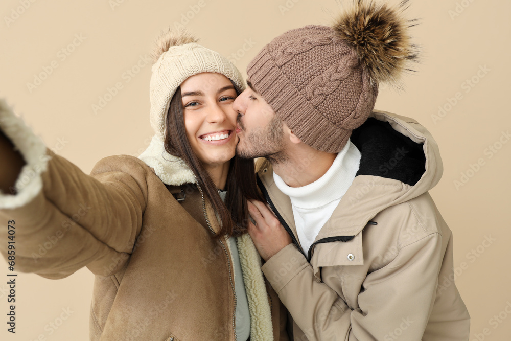 Loving couple in winter clothes taking selfie on beige background, closeup