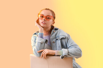 Poster - Beautiful young woman with stylish sunglasses sitting on chair against color background