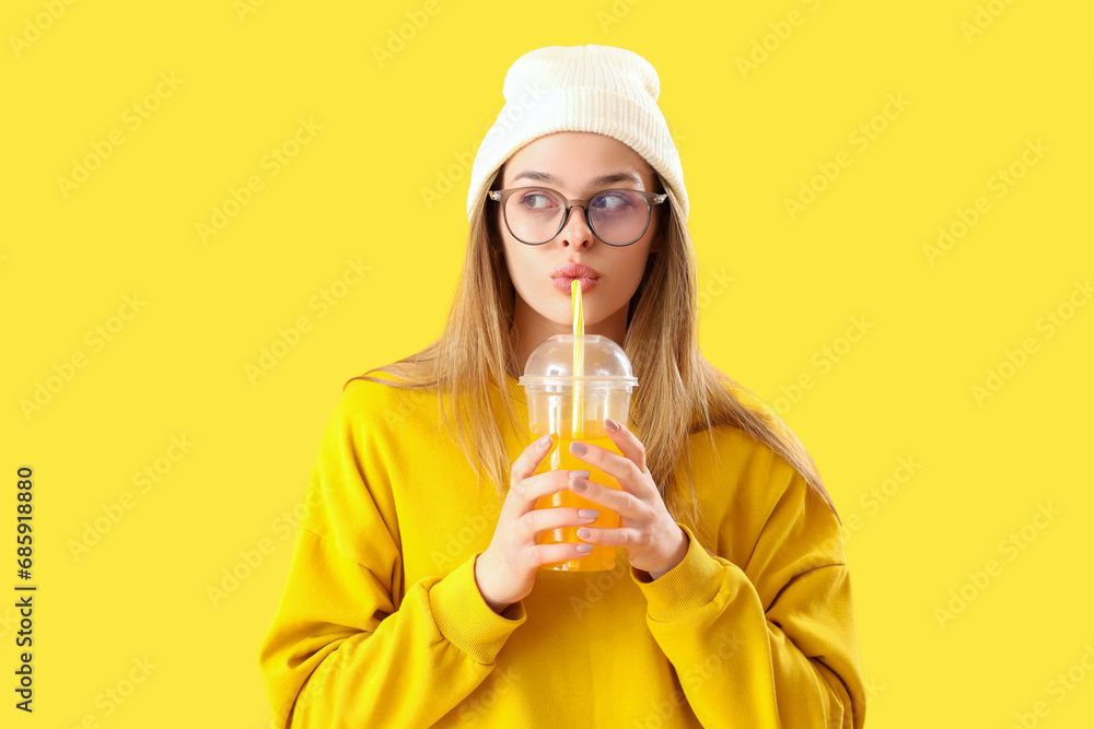 Young woman in eyeglasses drinking soda on yellow background