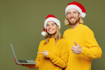 Wall Mural - Merry young couple two IT friend man woman wear sweater Santa hat posing hold use work on laptop pc computer show thumb up isolated on plain green background. Happy New Year Christmas holiday concept