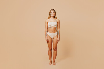 Full body smiling happy young nice lady woman with slim body perfect skin wear nude top bra lingerie stand looking camera isolated on plain pastel light beige background. Lifestyle diet fit concept.