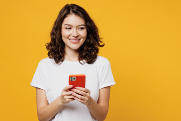 Wall Mural - Young smiling happy woman wear white blank t-shirt casual clothes hold in hand use mobile cell phone look aside on area isolated on plain yellow orange background studio portrait. Lifestyle concept.