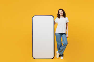 Wall Mural - Full body young smiling fun woman she wear white blank t-shirt casual clothes stand near big huge blank screen mobile cell phone smartphone with area isolated on plain yellow orange background studio.
