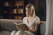 © nsit0108 - Blonde woman sitting on couch and reading a book. Female leisure reading time in living room. Generate ai