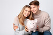© Ivan - Happy young couple smiling at camera holding big pink piggy bank isolated on pink background,theme of saving and putting money aside for home.generative ai