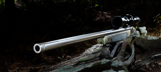 Rifle for hunting that uses black powder with a rifle scope resting on a log in a forest with barely any natural light coming through the trees. 