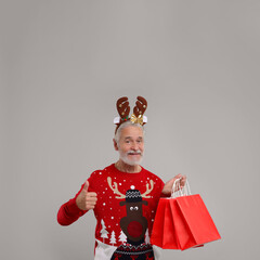 Sticker - Happy senior man in Christmas sweater and reindeer headband with shopping bags showing thumbs up on grey background