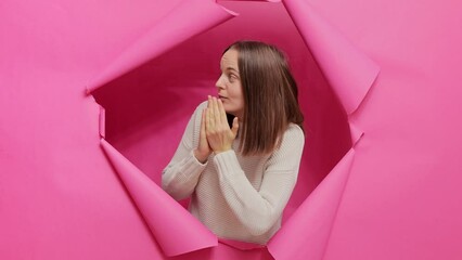 Wall Mural - Shocked brown haired woman wearing sweater posing in hole of pink paper wall looking with big eyes and covering open mouth sees advertisement.