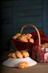 A wooden surface holds a bamboo basket filled with wheat cookies, juicy tutti frutti, and two jars of cherries side by side. High Resolution