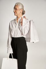 stylish middle aged woman in classy white shirt and black pants standing with shopping bags on grey