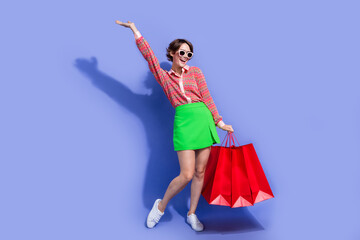 Sticker - Full size photo of positive woman dressed print shirt hold outfit in bags in sunglass raising up arm isolated on blue color background